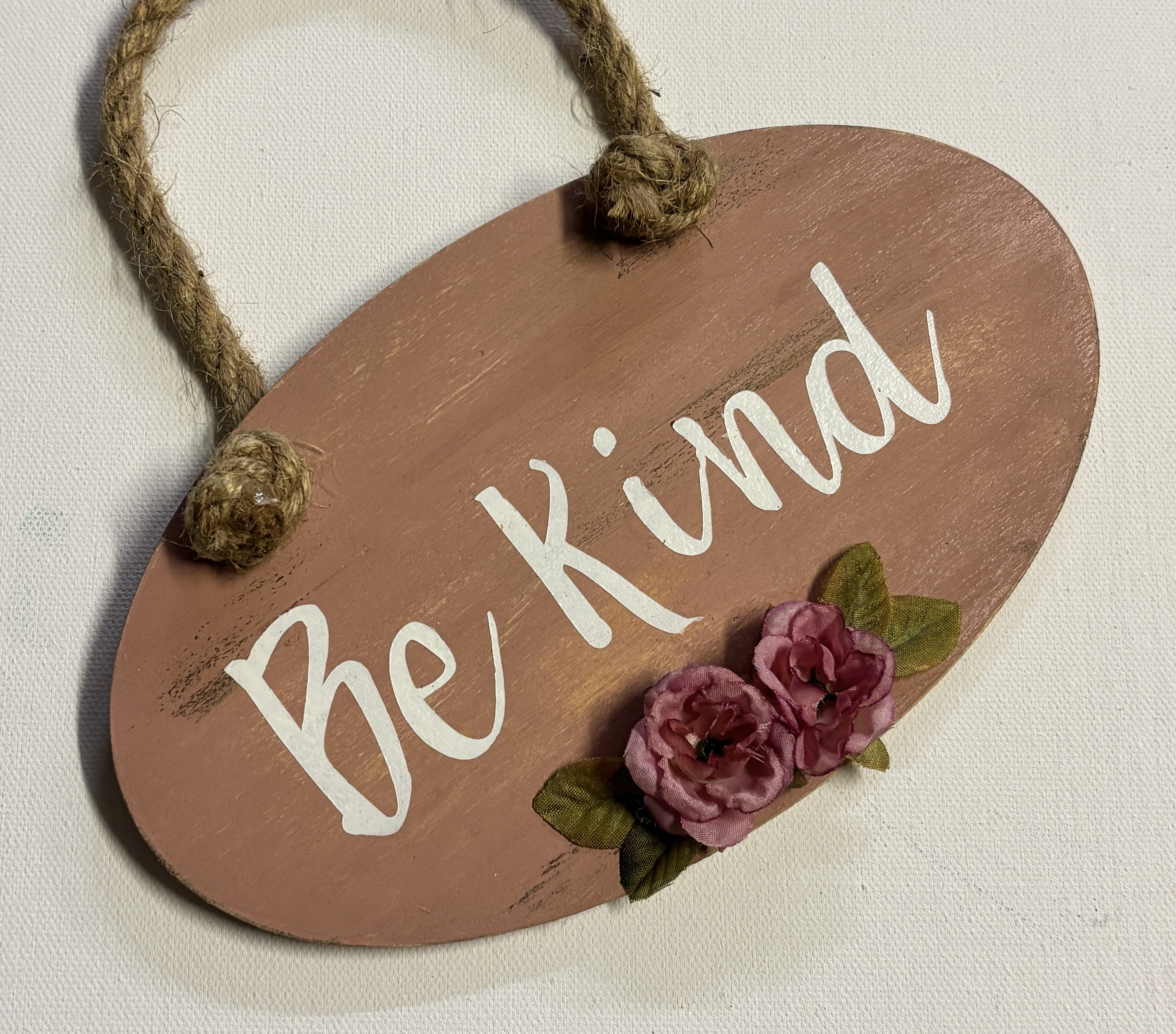 Terracotta oval wooden sign reading Be Kind with pink rose embellishments